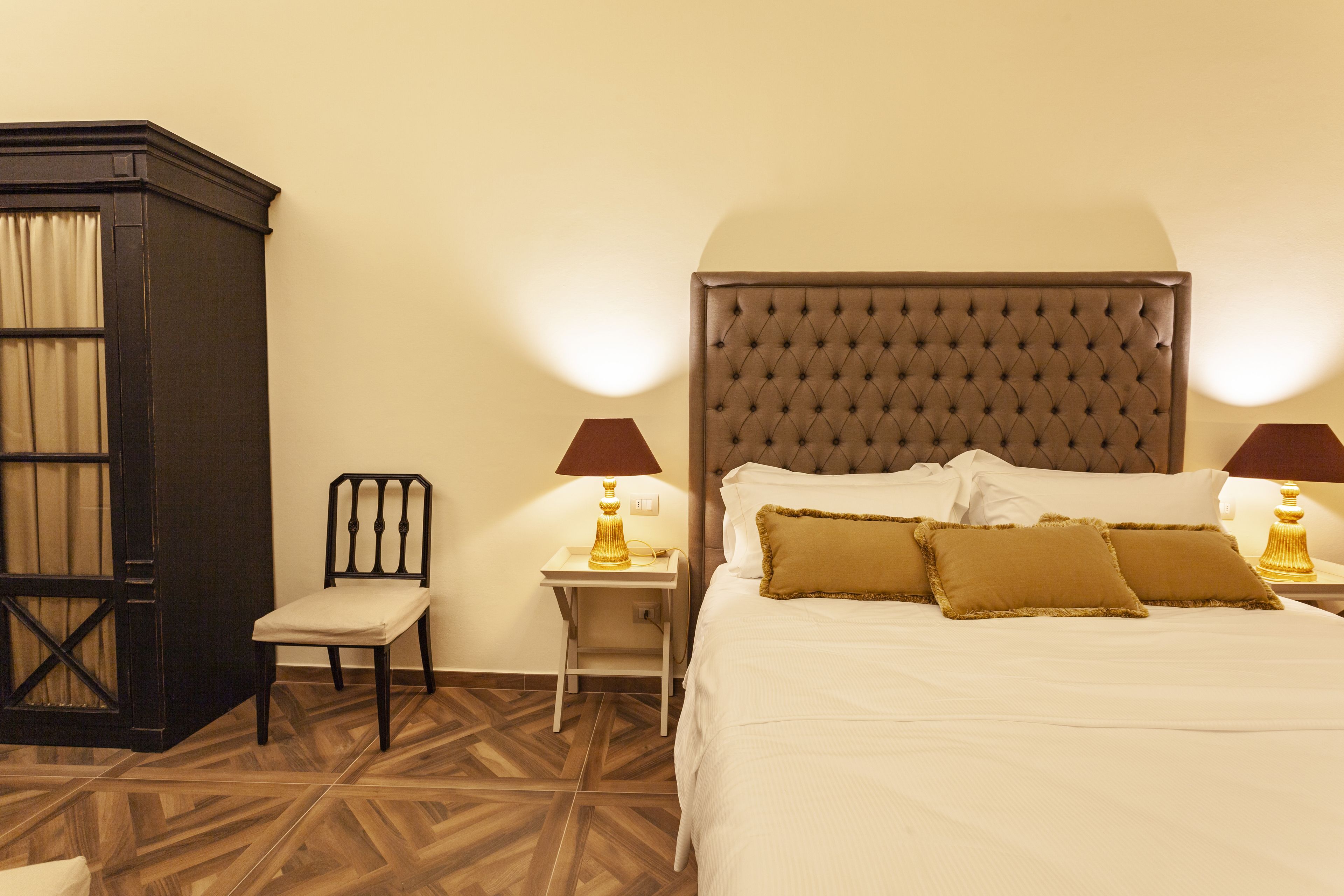 A hotel room with a large bed, two bedside tables with lamps, a wooden chair, and a dark-colored wardrobe.