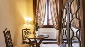Une pièce comprenant une table ronde, une chaise, un vase de fleurs, une lampe fixée au mur et une fenêtre avec des rideaux.