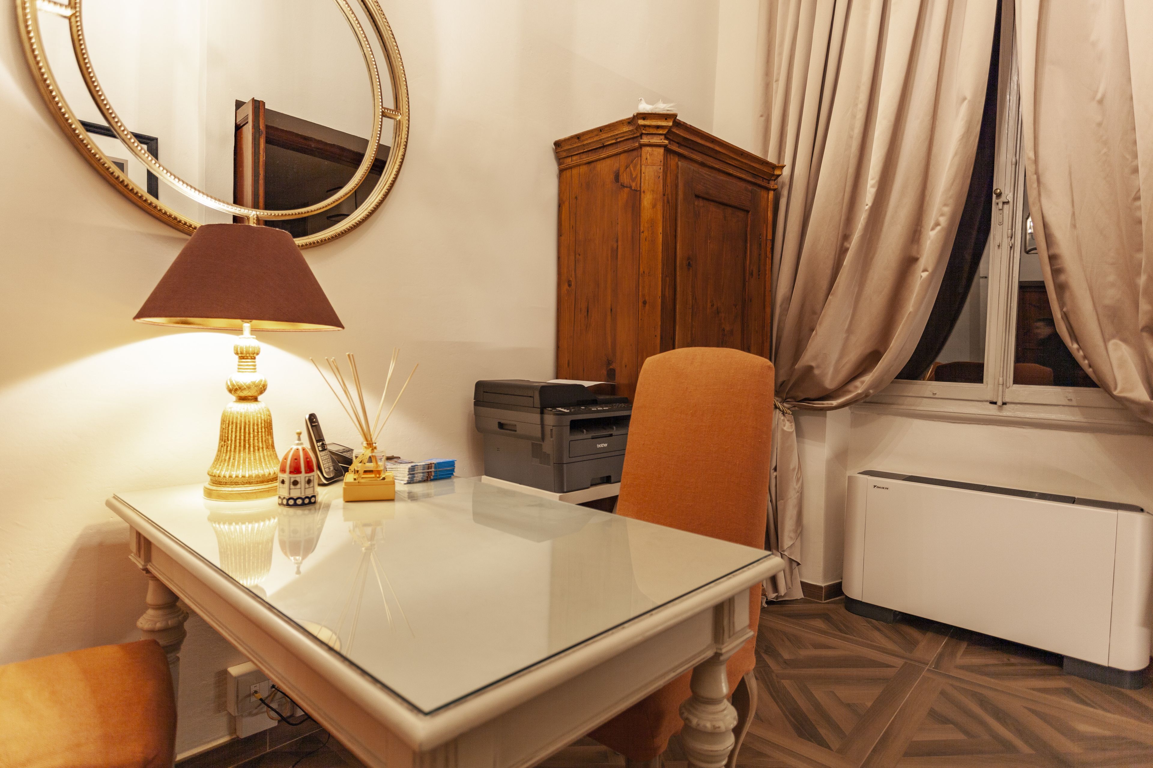 Ein Hotelzimmer mit Glastisch, Lampe, Drucker, Holzschrank und Heizkörper.