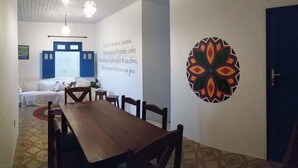 Un espace repas avec une longue table et des chaises en bois, un mur blanc orné d’un motif géométrique coloré et une porte bleue.