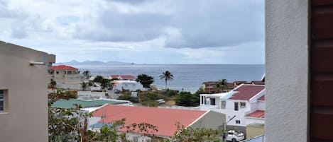 A coastal residential area with houses, a red-roofed building, and a view of the sea and mountains in the distance.