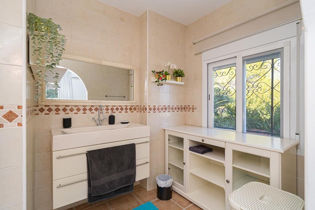 A bathroom with a large mirror, two sinks, a window with decorative iron bars, and a shelf with plants.