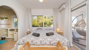 A bedroom with a bed, a wooden bench, a ceiling fan, a window with curtains, and a balcony with a wicker chair. - House in quiet and green surrounding where you can disconnect and enjoy family (La Nucia)