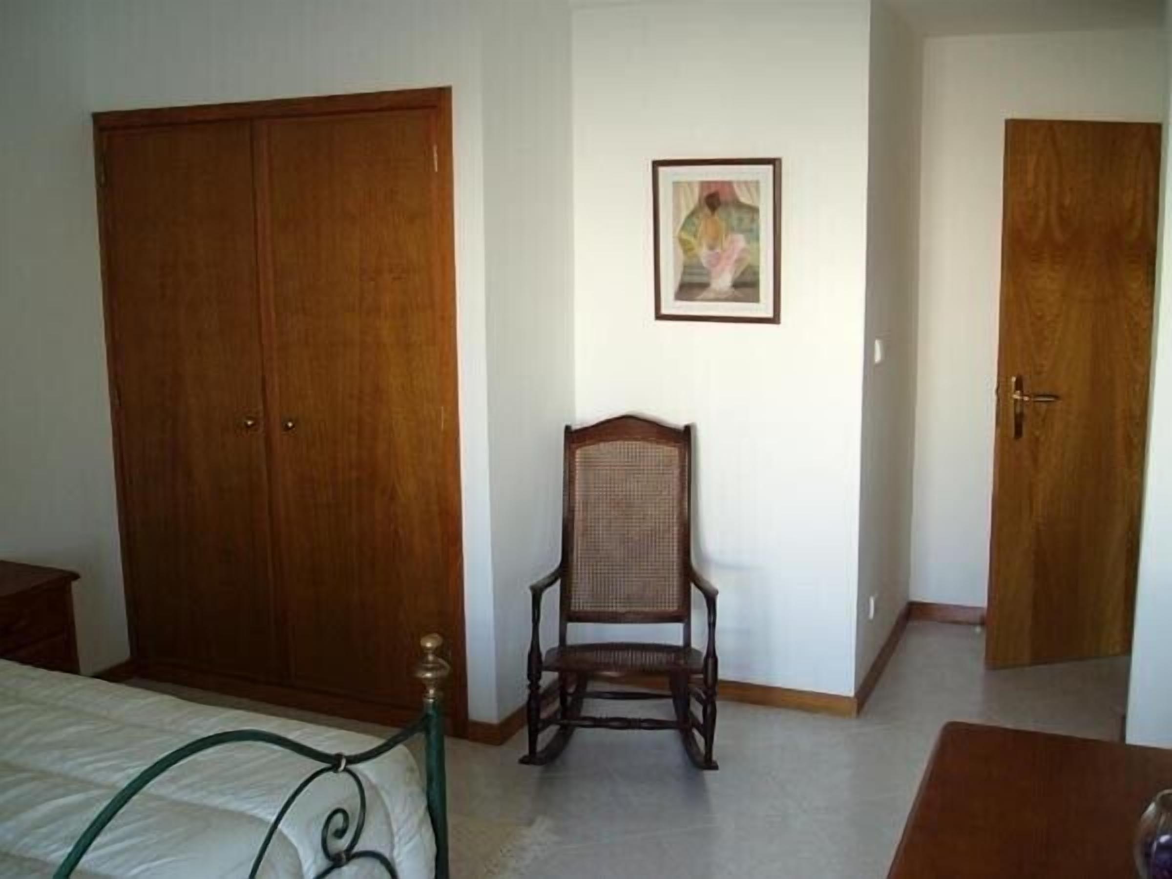 Ein Schlafzimmer mit großem Holzschrank, einem Korbstuhl, einem gerahmten Bild an der Wand und einem Bett mit grünem Metallgestell.