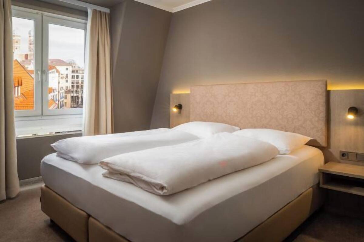 A double bed with a headboard, two bedside tables, a window with curtains, and a city view.