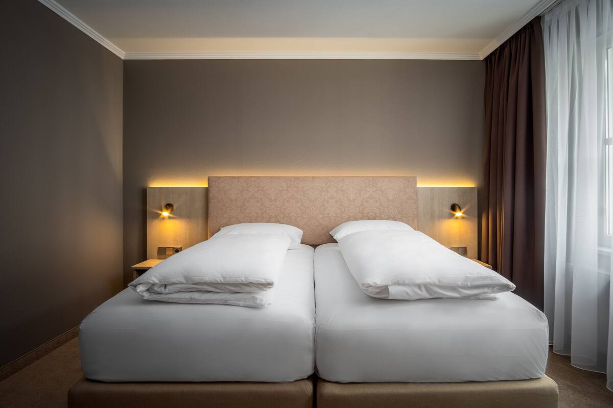 A double bed with white linens, two bedside tables with lamps, and a large window with sheer curtains.