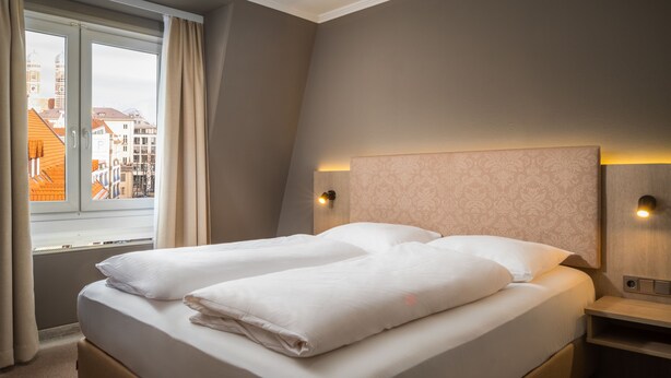 Habitació d'hotel amb un llit gran, dues taules de nit, una finestra amb cortines i vista a la ciutat.