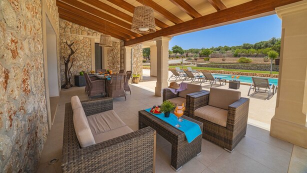 A covered outdoor seating area with wicker furniture, a pool in the background, and a stone wall.