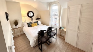 A bedroom with a bed, two chairs, a small table, a window with curtains, a mirror, and a shelf with decorative items. - Holland House (Wallasey)