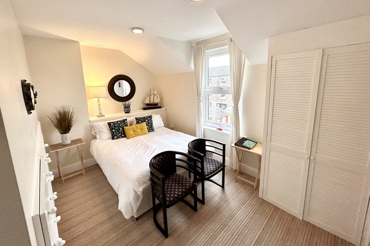 A bedroom with a bed, two chairs, a small table, a window with curtains, a mirror, and a shelf with decorative items.