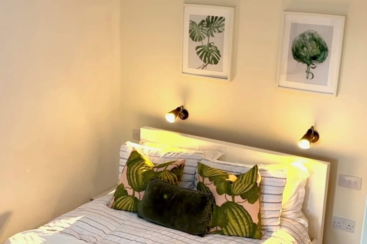 A bedroom with a bed, two wall-mounted light fixtures, and two framed botanical prints.