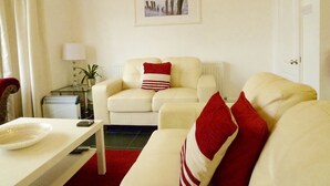 A living room with two cream sofas, a white coffee table, a red and white striped pillow, a framed picture on the wall, and a small side table with a lamp and a plant. - Holland House (Wallasey)