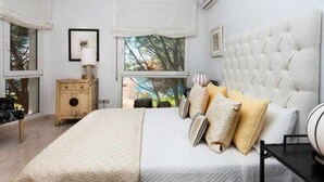 A bedroom with a large bed, a wooden nightstand, a bedside table, and a window with a view of trees. - VILLA LANTANA - Class and style with incredible views over Mahon harbour (Mao)