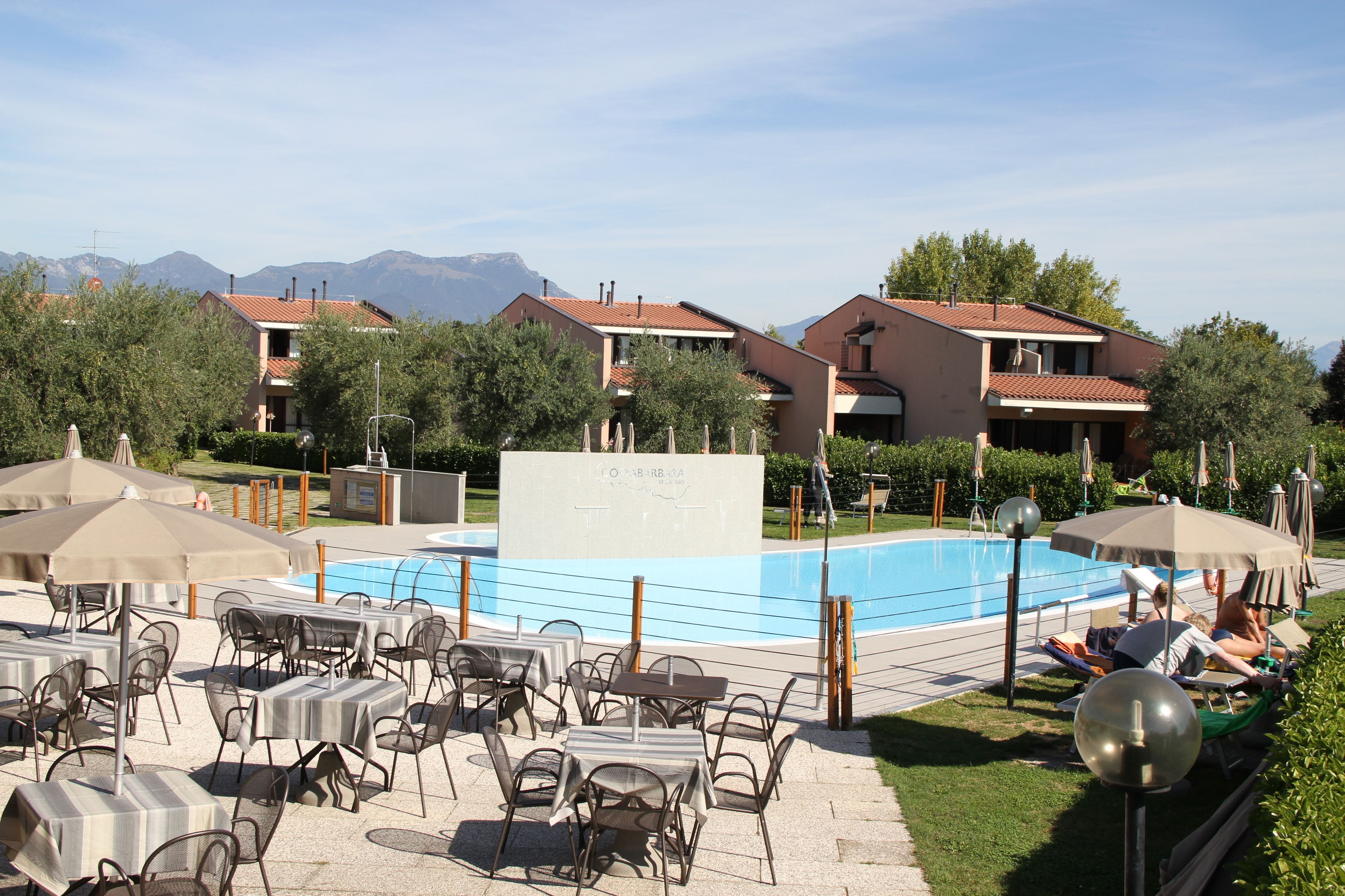 Un espace aménagé au bord de la piscine, avec des tables et des chaises, une piscine et des bâtiments en arrière-plan.