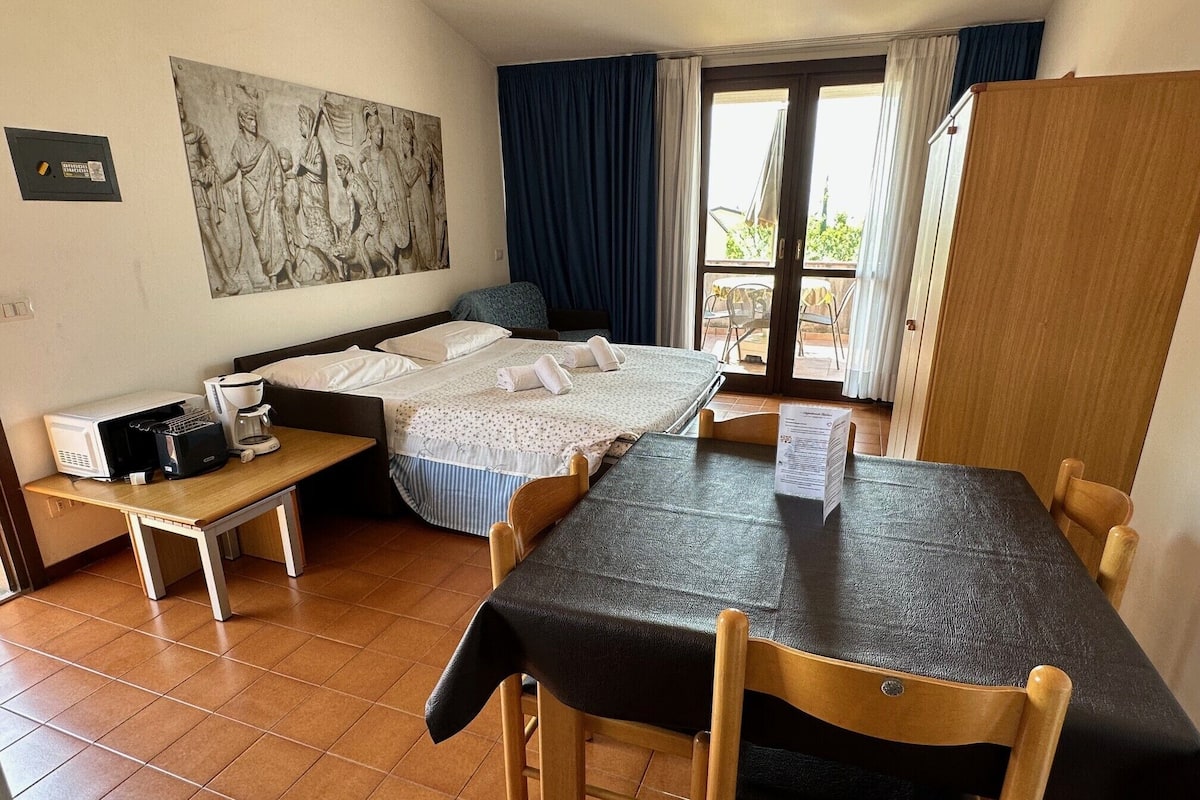 A hotel room with a bed, a desk with a coffee maker, a dining table, and a large window with curtains.