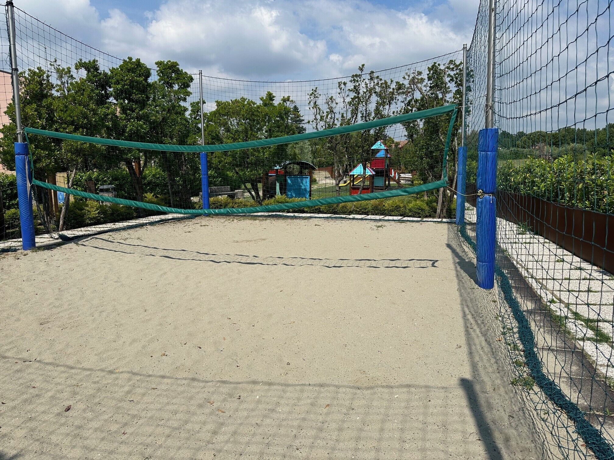 A sandy volleyball court enclosed by a net, surrounded by trees and a playground in the background.
