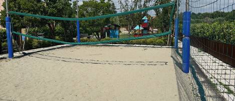 A sandy volleyball court enclosed by a net, surrounded by trees and a playground in the background.