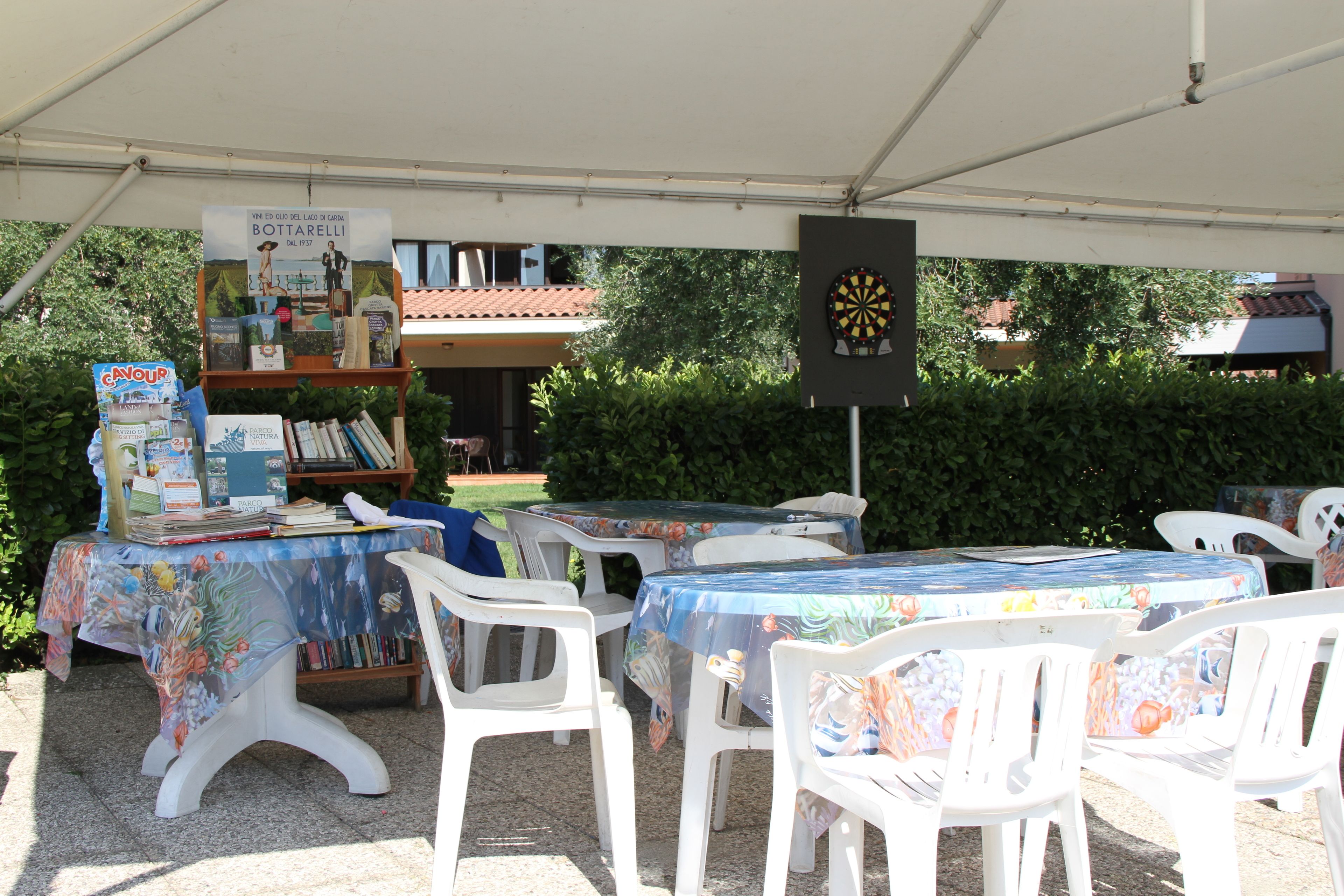 An outdoor market setup with tables covered in colorful tablecloths, white plastic chairs, and a dartboard.