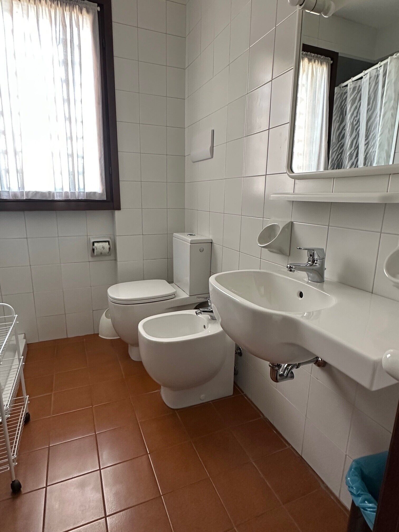 A bathroom with white tiles, a toilet, bidet, sink, and a window with curtains.