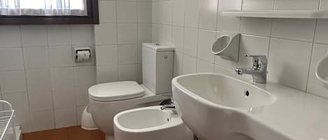 A bathroom with white tiles, a toilet, bidet, sink, and a window with curtains.
