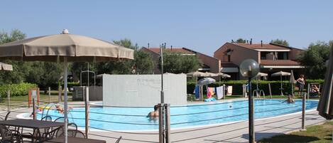 A swimming pool area with lounge chairs and umbrellas, surrounded by greenery and buildings.