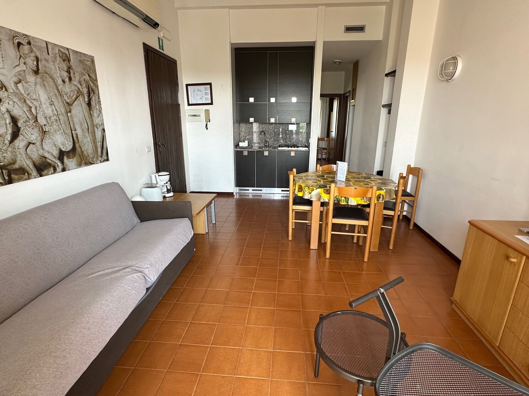 A living room with a sofa, a dining table with chairs, a kitchen area, and a door leading to another room.