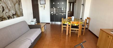 Sala de estar com sofá, mesa de jantar e cadeiras, espaço de cozinha e uma porta que dá para outro cômodo.