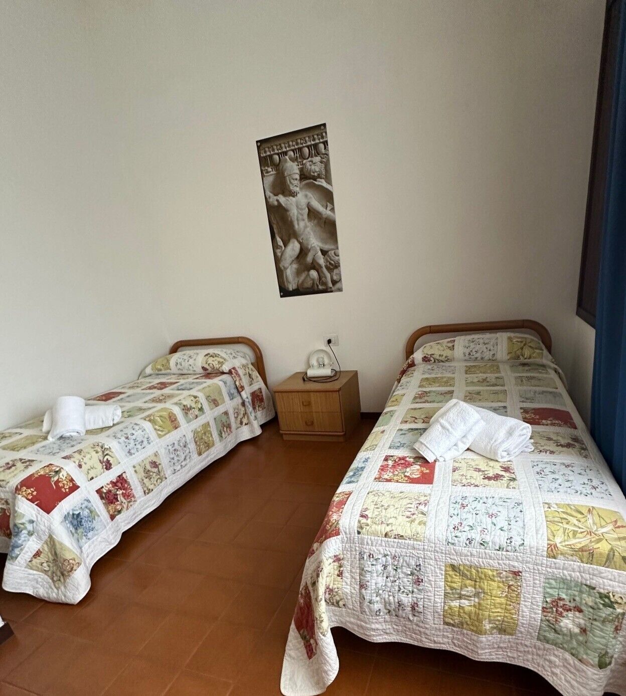 Ein Zimmer mit zwei Betten, jedes mit einer Blumensteppdecke und einem weißen Handtuch. Ein Nachttisch mit einer Lampe und einem Poster befindet sich zwischen den Betten.