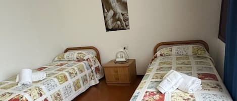 Ein Zimmer mit zwei Betten, jedes mit einer Blumensteppdecke und einem weißen Handtuch. Ein Nachttisch mit einer Lampe und einem Poster befindet sich zwischen den Betten.
