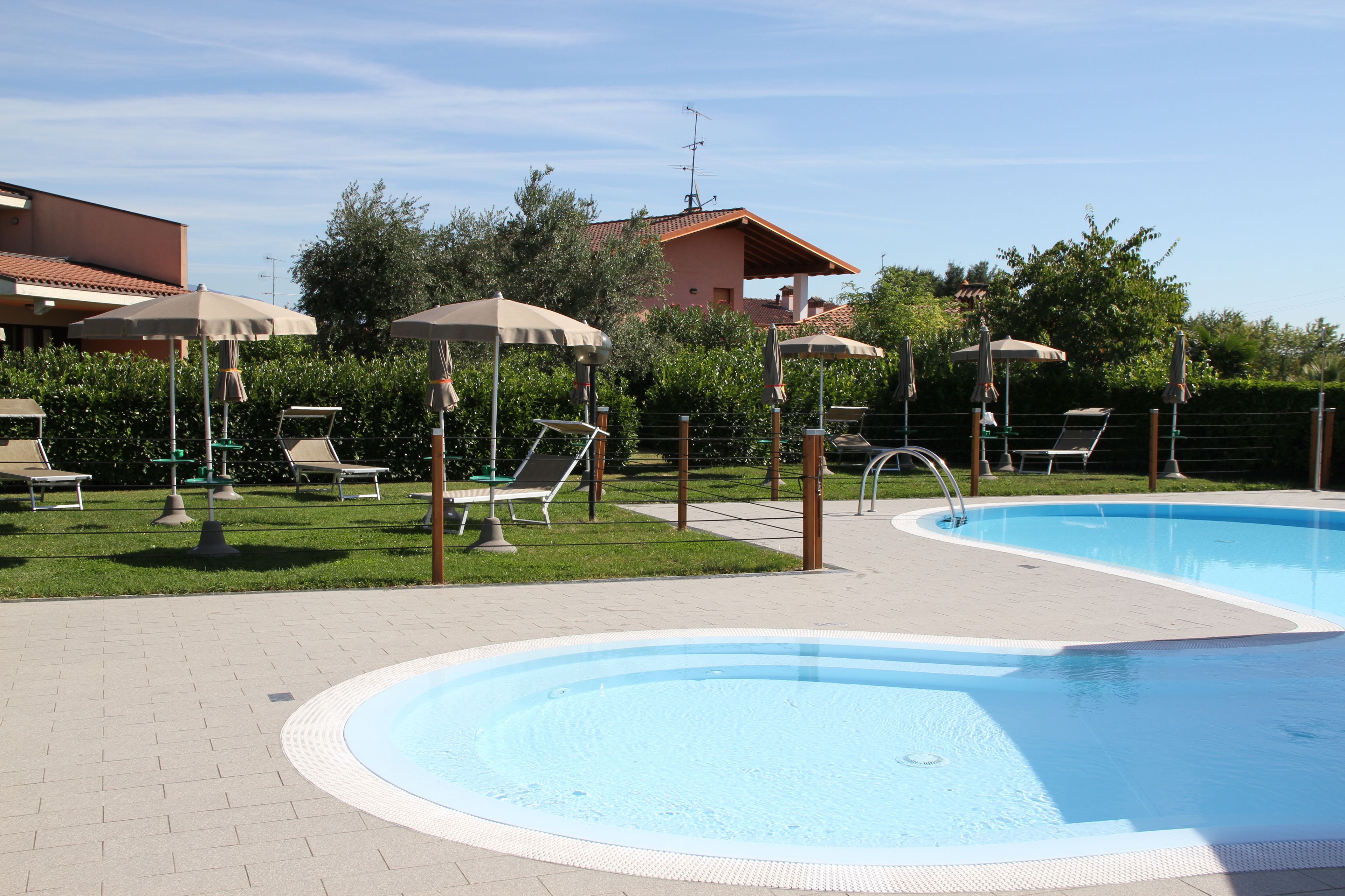 Una piscina con sillas de descanso y sombrillas, rodeada de vegetación y un edificio.