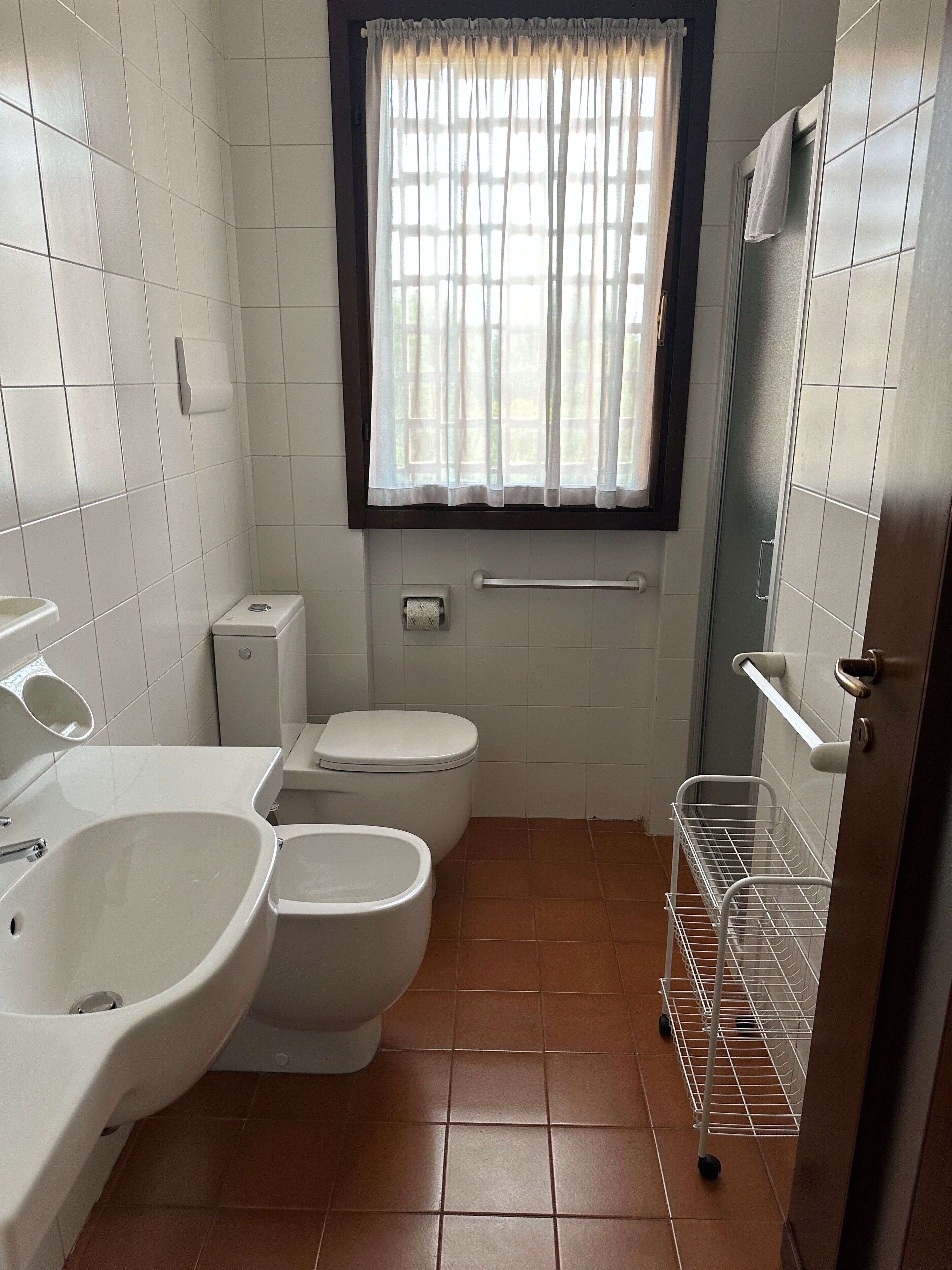 Een kleine badkamer met toilet, bidet en wastafel. Er is een raam met gordijnen, een handdoekhouder en een metalen plank.