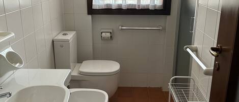 Une petite salle de bain avec des toilettes, un bidet et un lavabo. On y trouve une fenêtre avec des rideaux, un porte-serviettes et une étagère en métal.