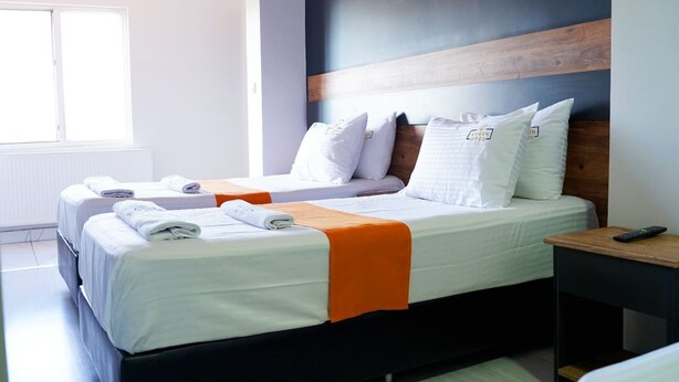 A hotel room with two beds, each with white bedding and orange throw blankets. There is a wooden headboard, a nightstand with a remote control, and a window providing natural light. - Ankara Otel Evren (Ankara)