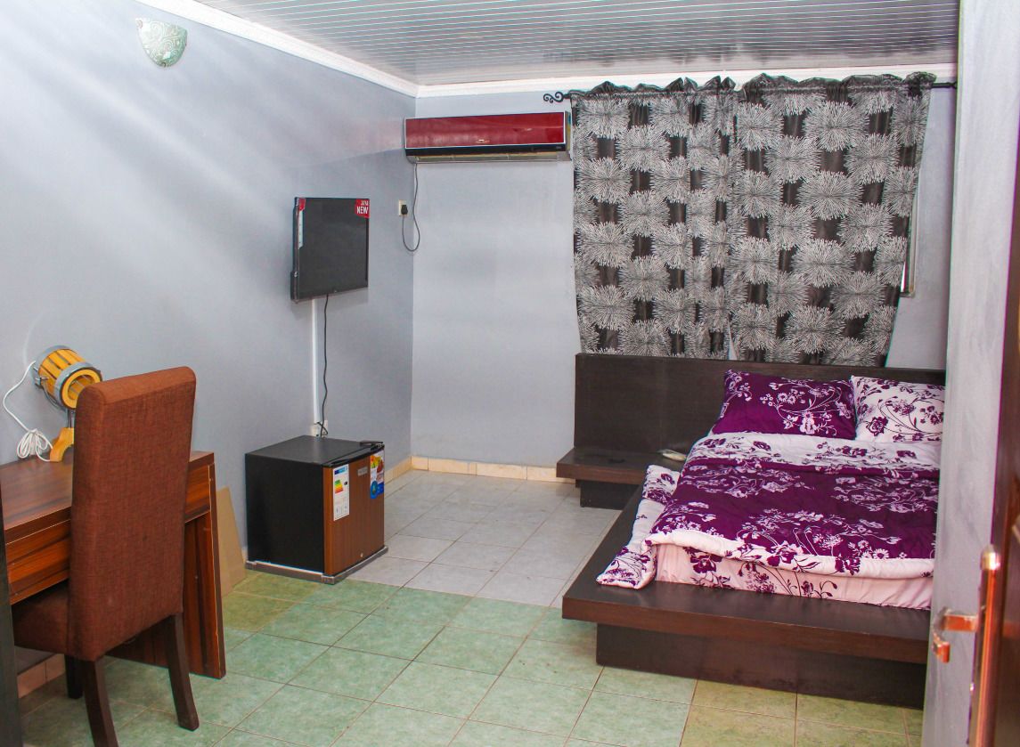 A compact hotel room with a bed, a desk, a chair, a TV, and an air conditioning unit.