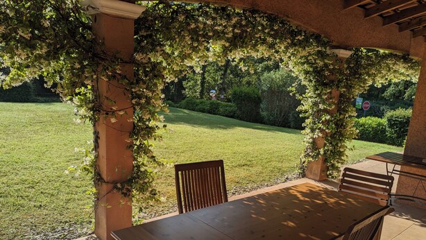 A covered patio with a wooden table and chairs, surrounded by greenery and a view of a lawn.