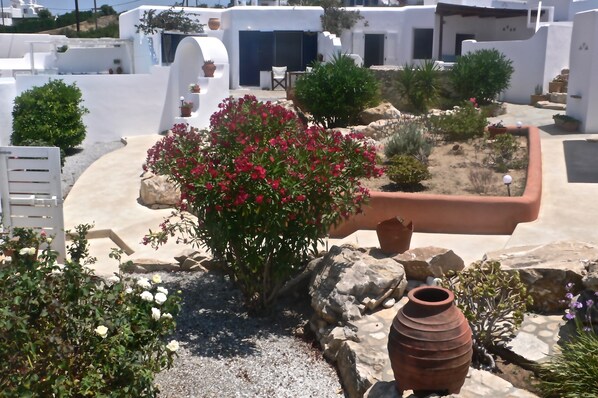 A white-washed building with a garden featuring a large bush with pink flowers, a rustic pot, and a small white chair.