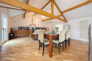 Un vaste espace à manger avec une table et des chaises en bois, une cuisine à l’arrière-plan et un plafond haut avec des poutres apparentes.