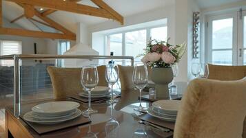 A dining table set with plates, glasses, and a vase of flowers, in a room with wooden beams and large windows.