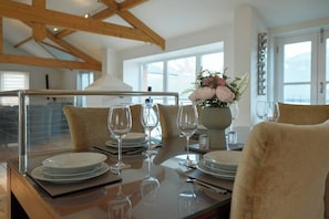 A dining table set with plates, glasses, and a vase of flowers, in a room with wooden beams and large windows.