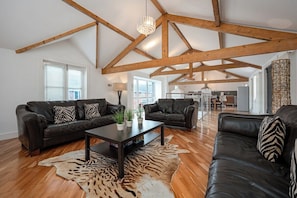 A spacious living room with high ceilings and exposed wooden beams, furnished with leather sofas and a coffee table, and a large window providing natural light.