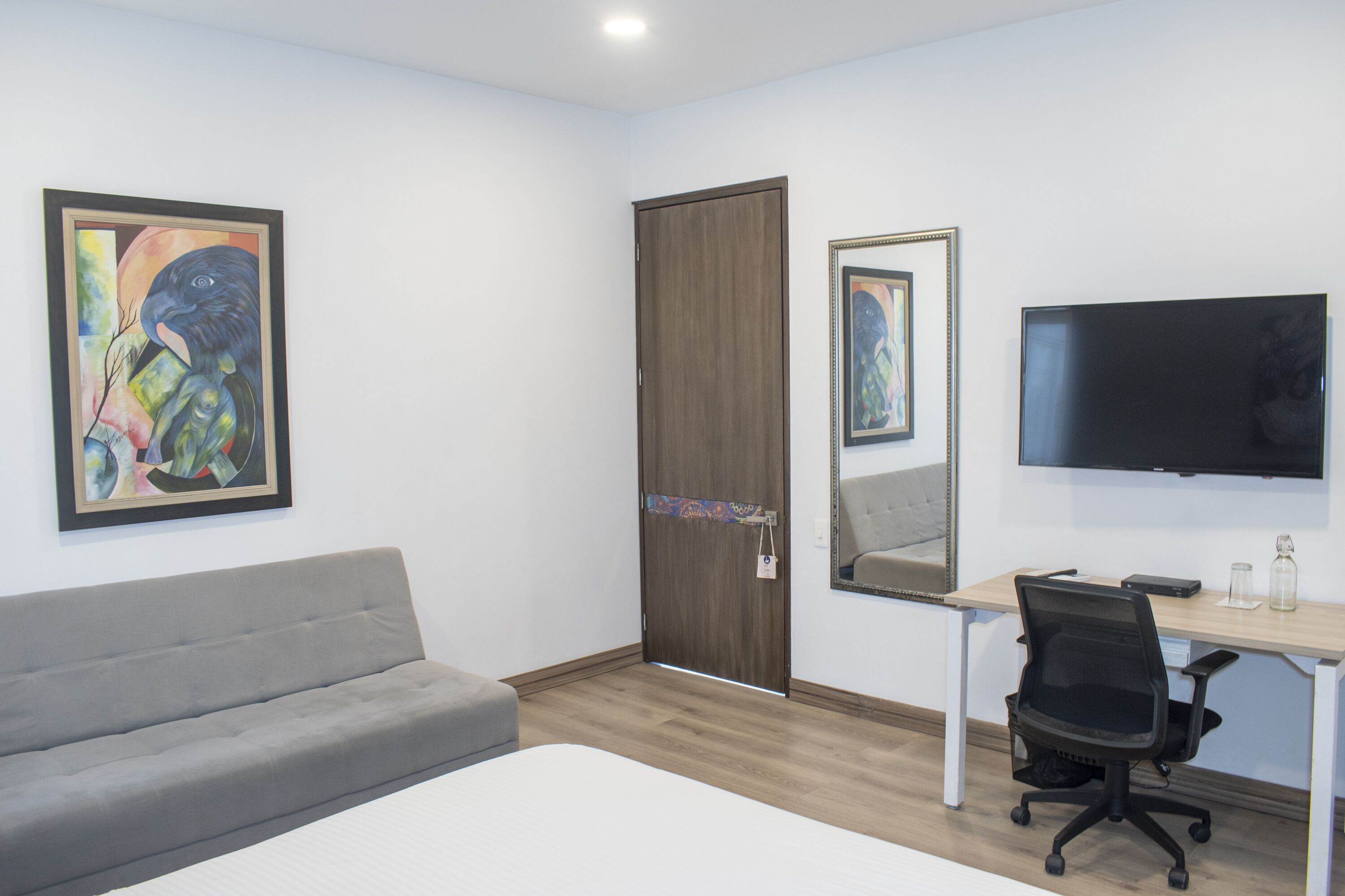 A room with a grey sofa, a wooden door, a desk with a chair, and a flat-screen TV mounted on the wall.