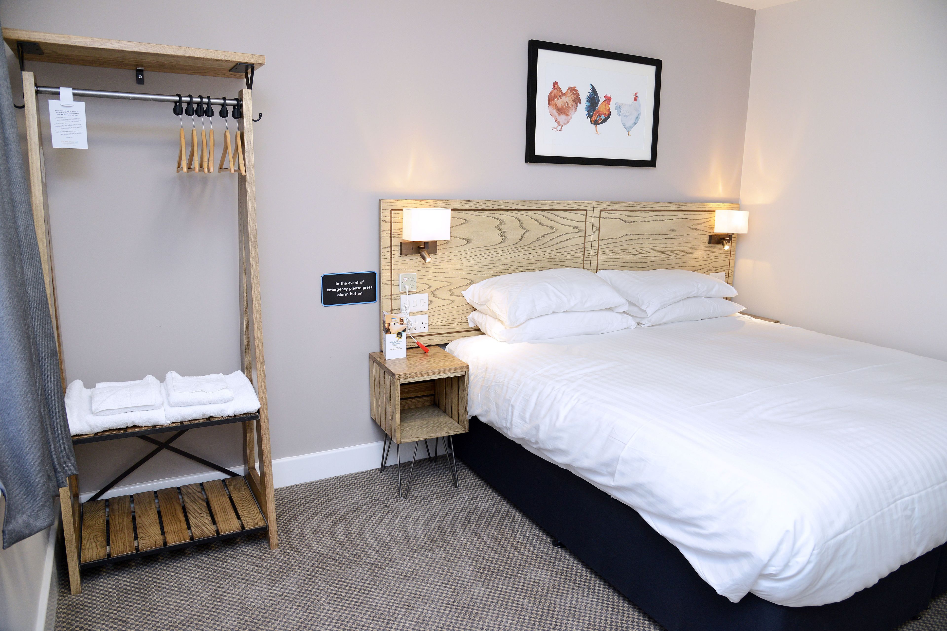 A hotel room with a bed, a wooden nightstand with a lamp, a wooden shelf with towels, and a framed picture on the wall.