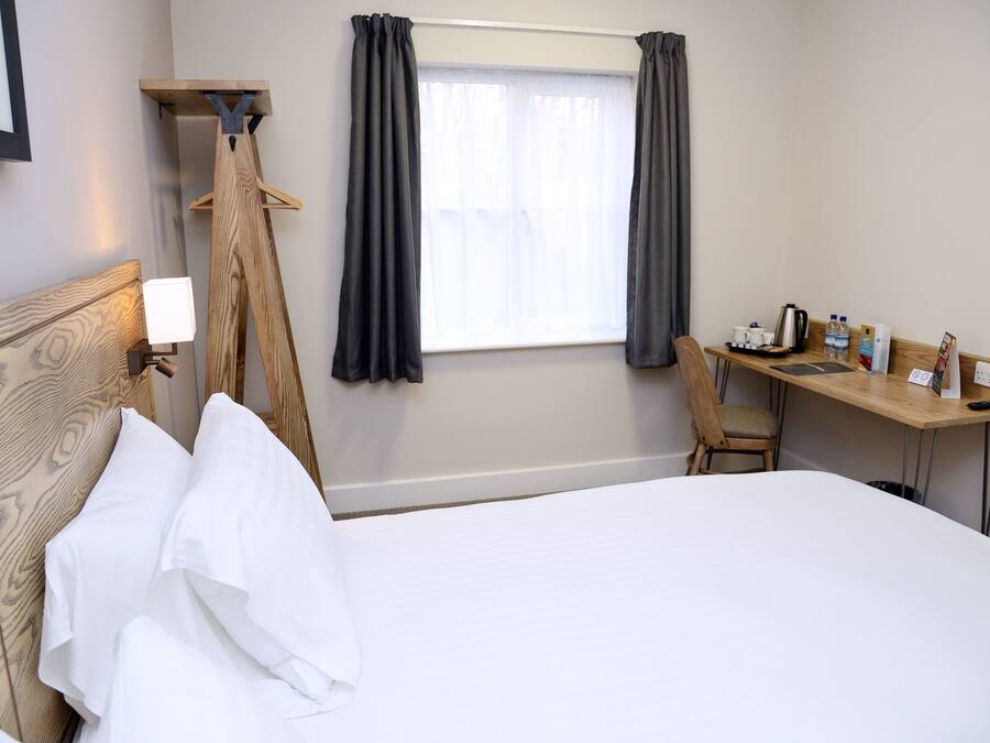 Habitación de hotel con cama, escritorio, silla, perchero y ventana con cortinas.