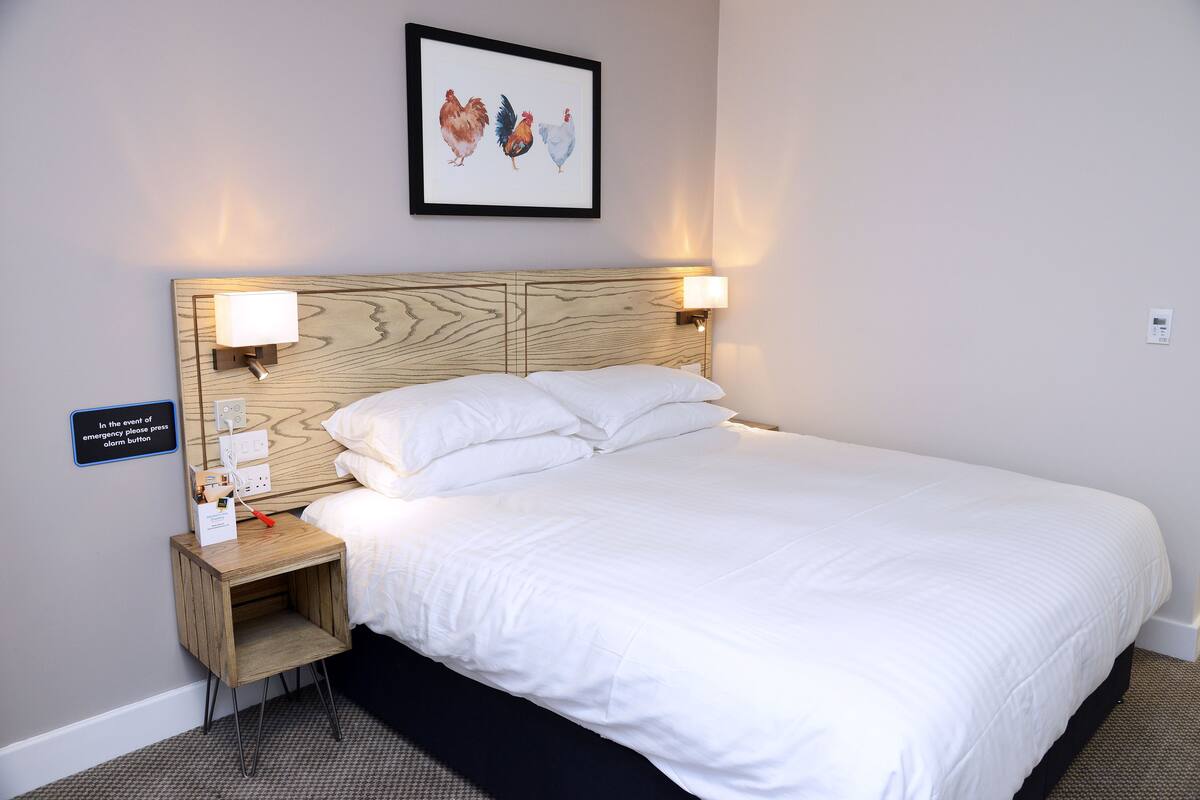 A neatly made bed with white linens, a wooden headboard, two bedside lamps, a small wooden nightstand with a charging station, and a framed artwork of chickens on the wall.