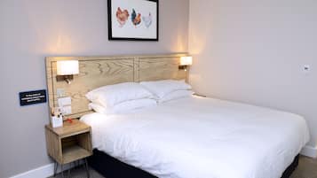 Een netjes opgemaakt bed met witte lakens, een houten hoofdbord, twee bedlampen, een klein houten nachtkastje met een oplaadstation, en een lijst met daarin een afbeelding van kippen aan de muur.