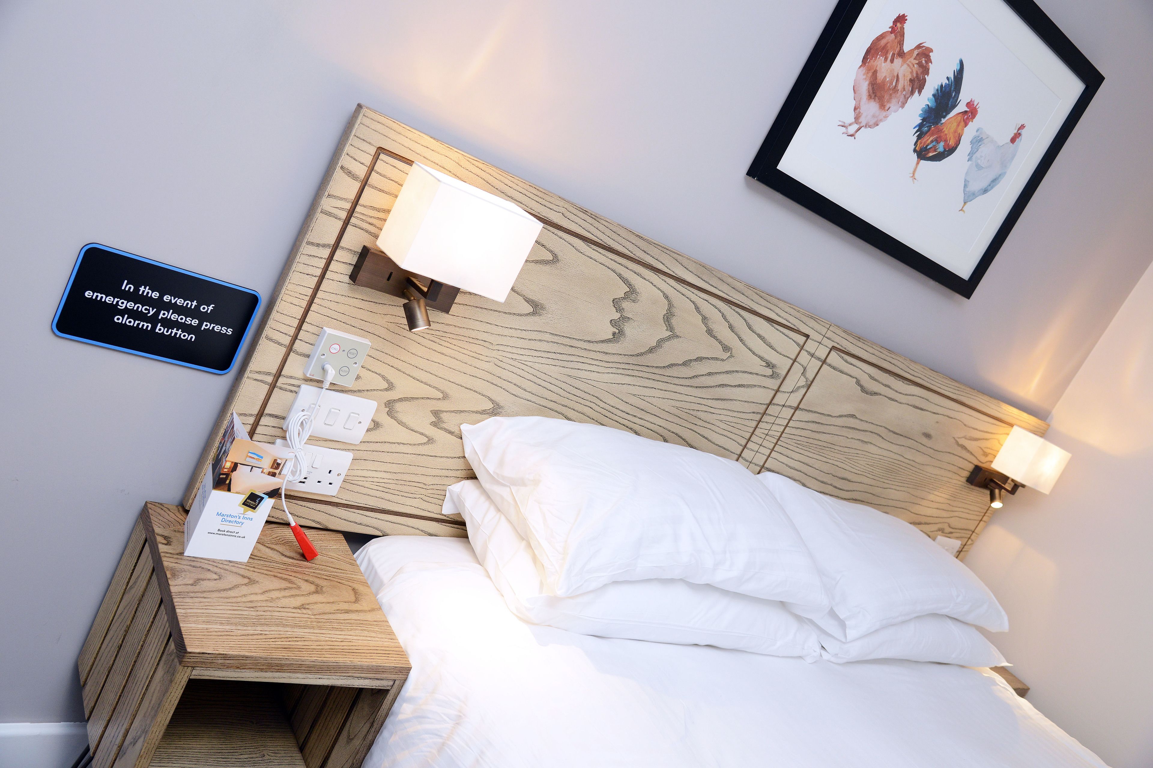 Habitación de hotel con cabecera de madera, ropa de cama blanca, mesita de noche con lámpara, un cuadro enmarcado con gallinas y una señal de alarma de emergencia.