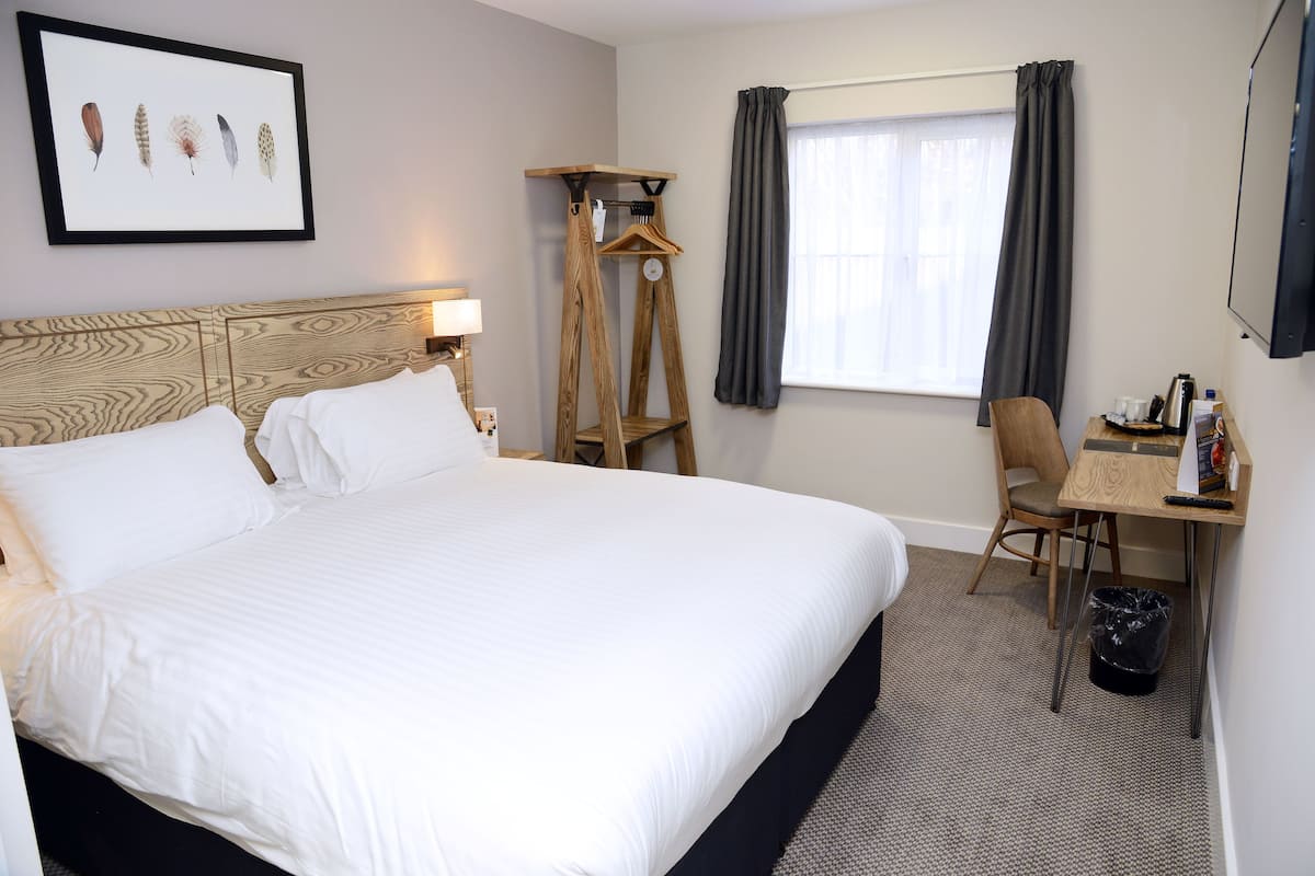 A hotel room with a large bed, a desk with a chair, a coat rack, a clock, and a framed picture on the wall.