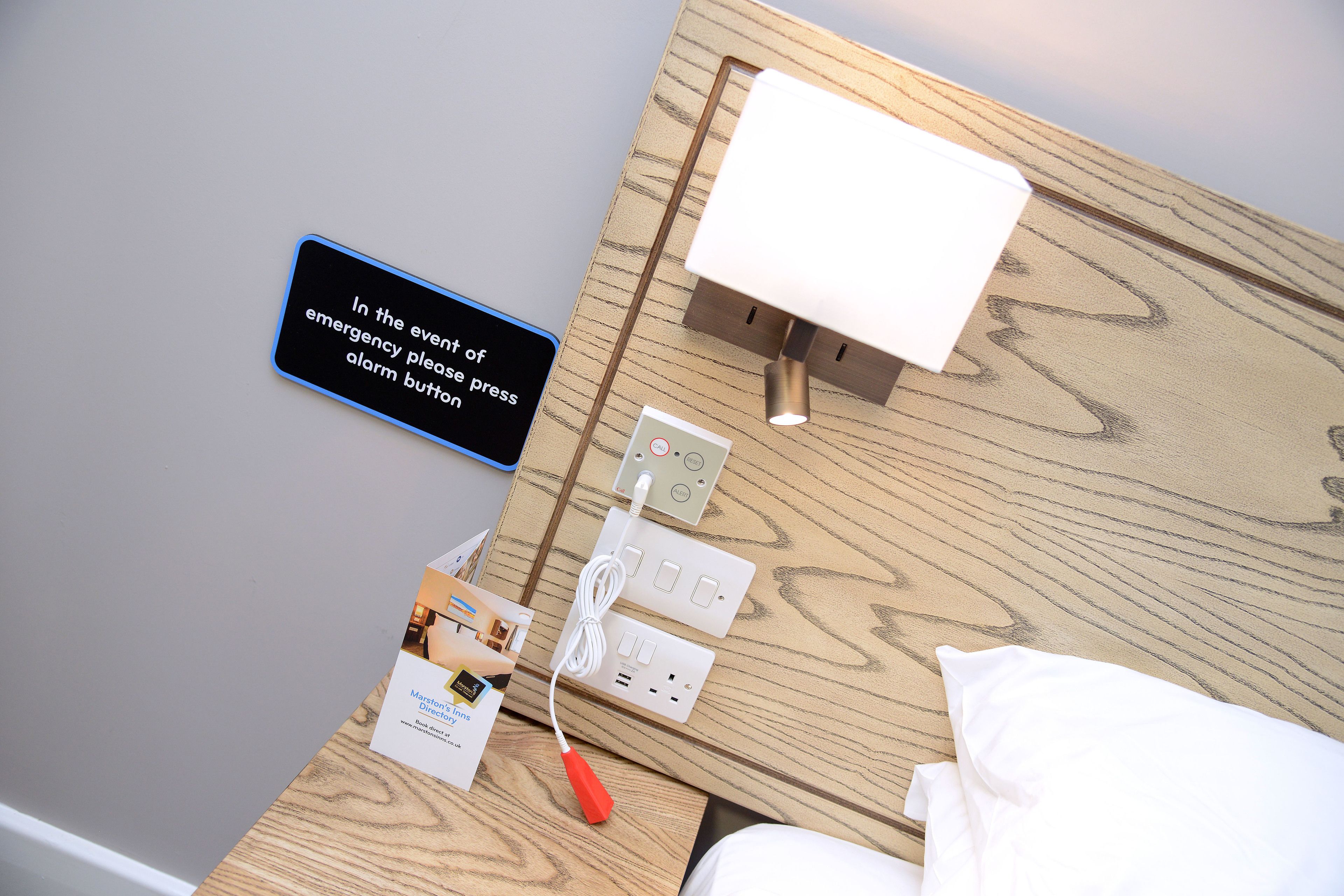 Une table de chevet en bois, sur laquelle se trouvent une lampe blanche, une multiprise et un panneau indiquant qu’il faut appuyer sur le bouton d’alarme en cas d’urgence.