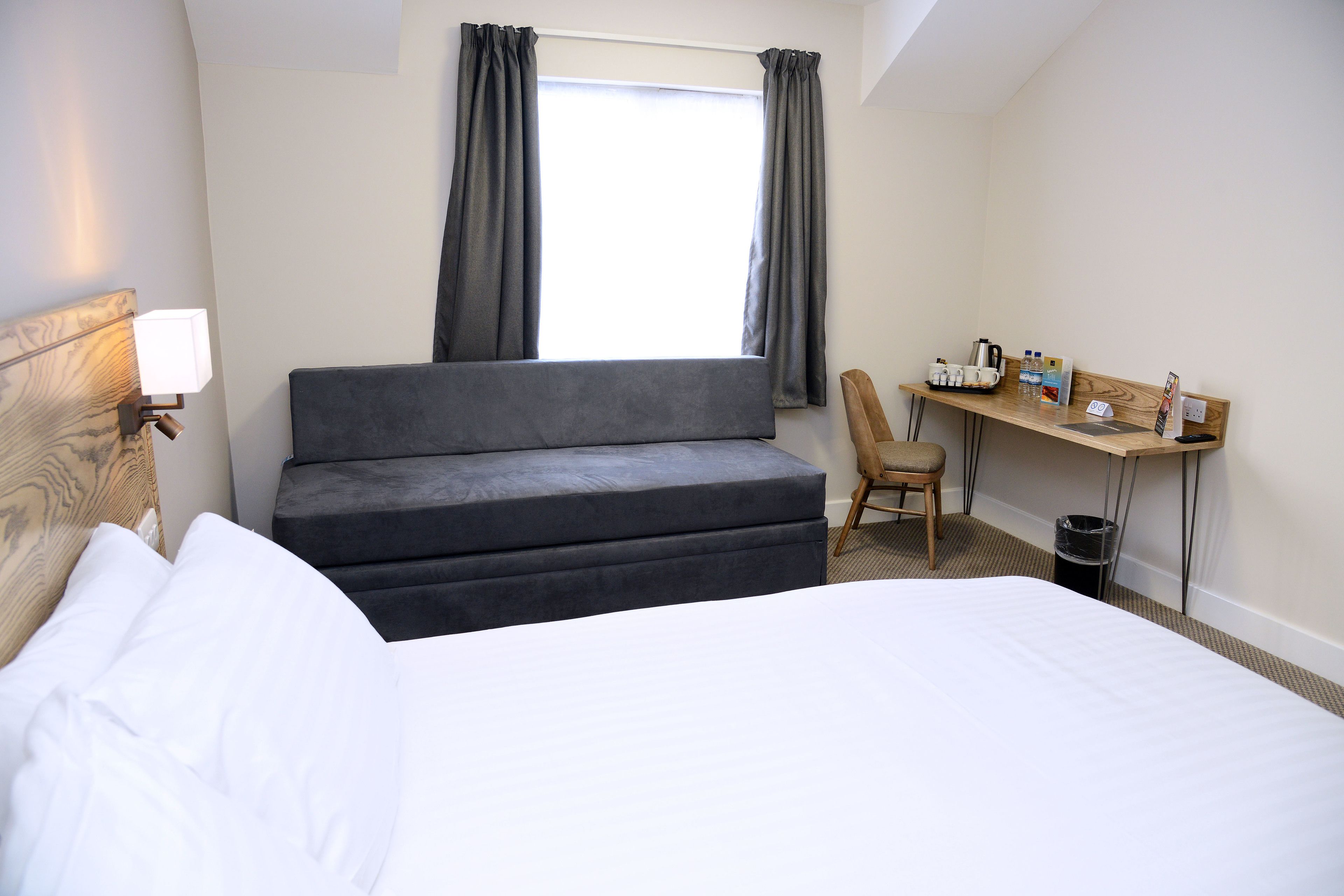 A hotel room with a bed, a sofa, a desk with a chair, a window with curtains, and a nightstand with a lamp.
