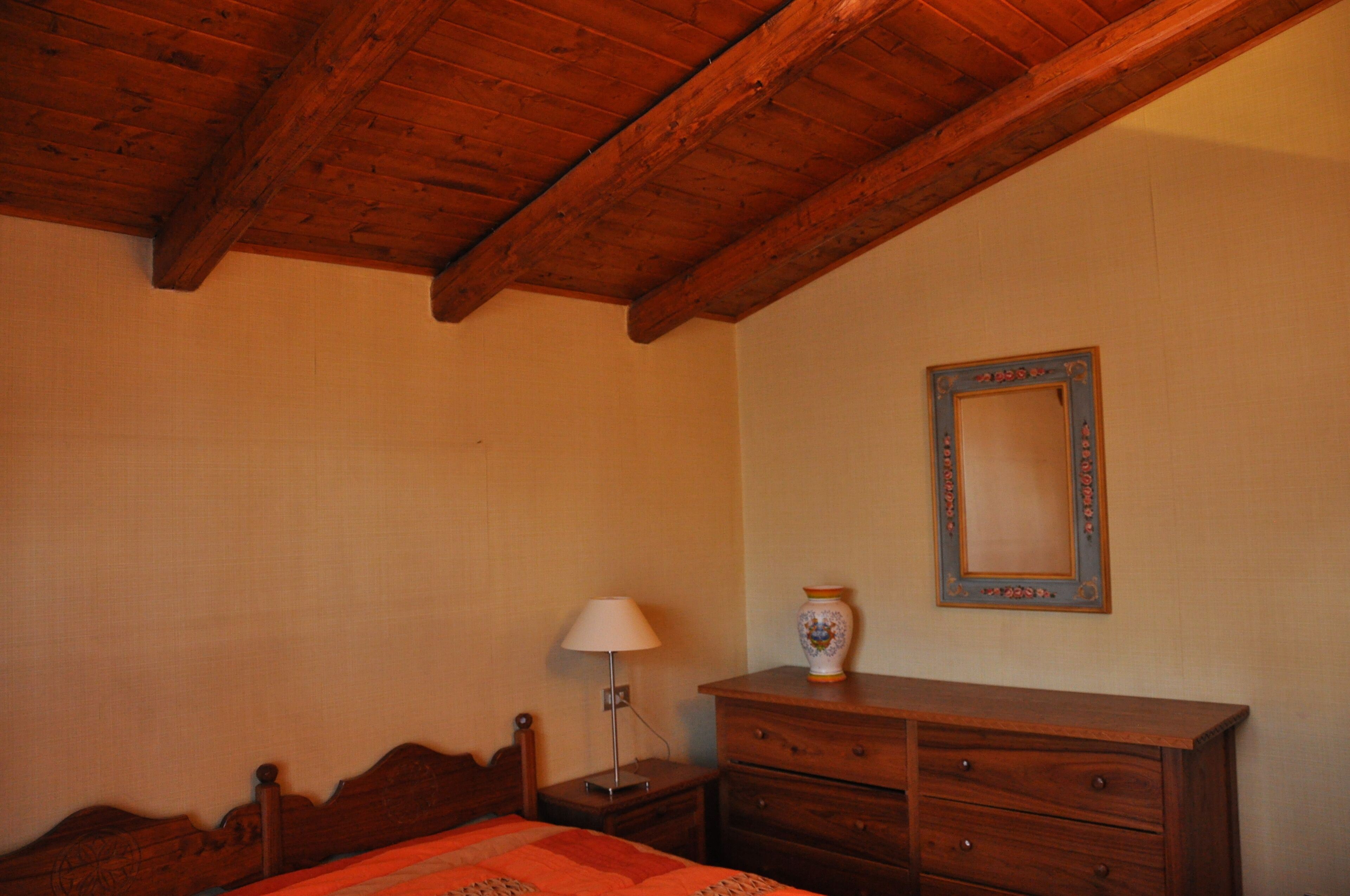 A bedroom with a wooden ceiling, a bed, a nightstand with a lamp, a dresser, and a mirror on the wall.